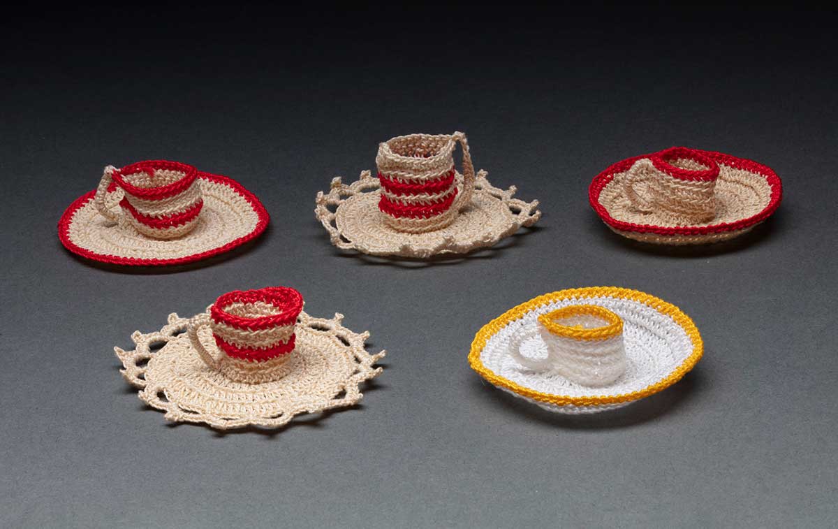 A set of five miniature crocheted teacups and saucers. Two are made with beige coloured thread with a red rim on the cup and saucer. Two also have beige thread and red stripes on the teacups, and the saucers have beige scalloped edges. One is made from white thread with a yellow rim on the cup and saucer. They are each approximately 3-4 centimetres in diameter. - click to view larger image