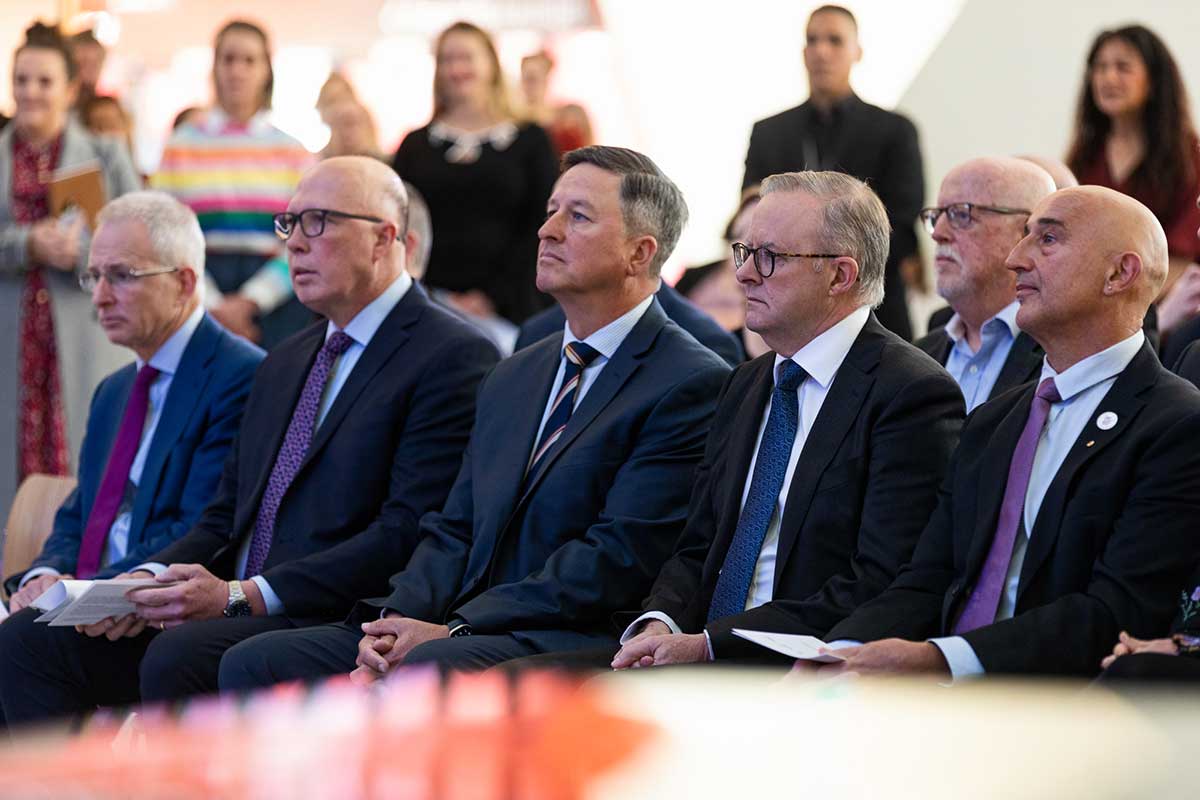 Five men sitting at an event, with people standing in the background.