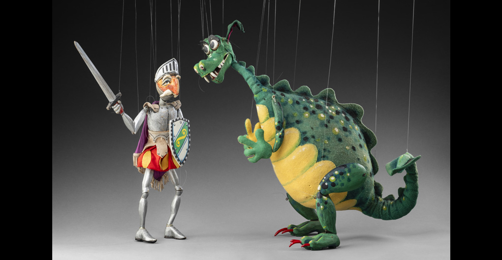 Two string puppets. Left is a puppet of a knight called Sir Giles. He has a white moustache and is wearing silver armour, red and yellow pants, and a purple and silver cape. He is holding a sword in one hand, the other holds a shield decorated in black, white, green, yellow and blue. The right puppet is a smiling dragon. Dragon is bigger than the knight. The dragon is painted green with a yellow stomach and red nails.