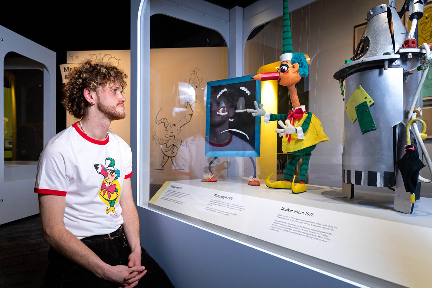 Side view of a man gazing at Blackboard, Squiggle and Rocket puppets in a display case at a museum. 