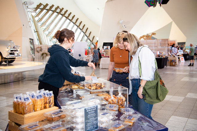 A woman serves a bag of baked goods to two women, browsing at a market stall. - click to view larger image