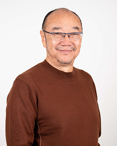 Photograph of Tony Ayres. Tony is wearing a brown shirt and glasses. He is smiling at the camera. 