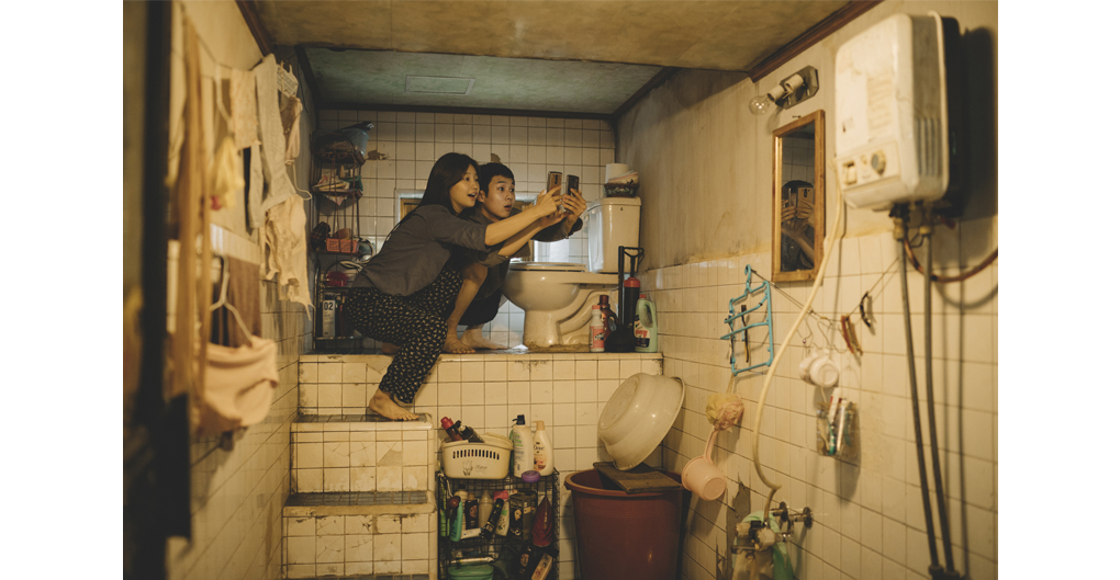 Film still of the bathroom set in the film Parasite. The bathroom is small and cramped, packed with an assortment of household items, and a toilet is raised on a platform. Two related characters, Kim Ki-jung (Jessica) and Kim Ki-woon (Kevin) are perched close together on the platform, holding out mobile phones, seeking wifi from a neighbour's property. The tiles are stained and aged, appearing neglecte and unhygienic. The scene is cramped, claustrophobic, dingy.