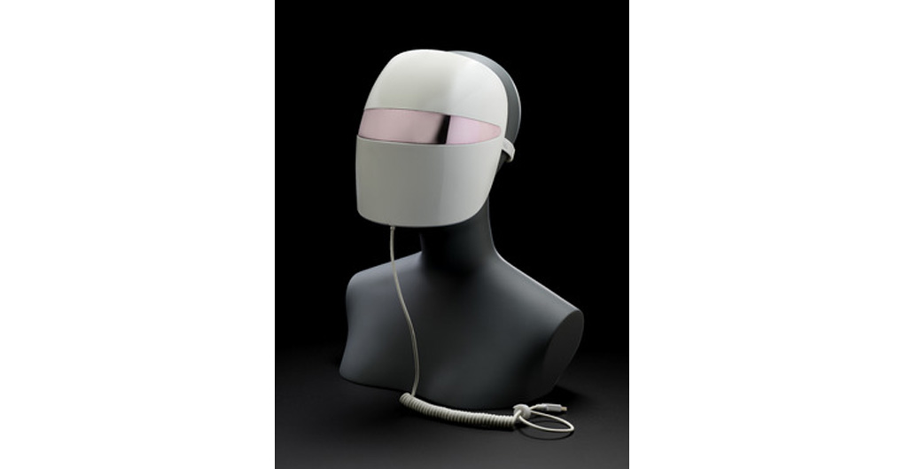 A circular shaped mask in a shiny light grey plastic, with a metallic eye strip and a single cord which ends in a long spiral. The mask is placed on a mannequin' torso head, which it wraps around, fully concealing the face, with a slight overhang over the chin.