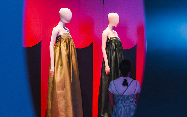 Photograph of a person looking at mannequins in the Museum's Hallyu exhibition. The woman is wearing a blue and white dress and has her hair pulled back. The photograph is taken looking through an arched shaped hole in exhibitions wall. The dresses on the mannequins are bold colours. Image credit: Tim Ngo.