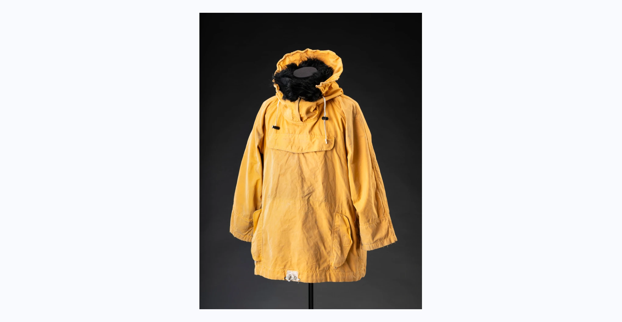  A yellow cotton Ventile jacket with a wired hood that has black fur, a zipper and drawstrings. It has a zip-up pocket with a velcro closure at the centre of the chest, zip-up access points on the left and right side, and smaller zip-up pockets with flaps and velcro closures on the lower left and right side. The interior of the jacket is lined with green cotton fabric. Sewn to the back of the neck is a cloth label with black text reading 'KINDRED MANUFACTURING / PTY. LTD. / SIZE SM / MADE IN AUSTRALIA / NAME'.