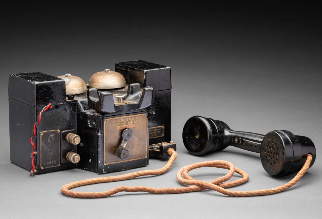 A black, hand cranked telephone set. It consists of a metal box with two metal bells on top and a black metal and plastic turnable handle on the front. Behind this handle is a metal plaque that reads 'REPLACE HANDSET / WHEN FINISHED / AND RING OFF / TELEPHONE SET F MK. 1*'. Impressed into the metal below the plaque is '939'. Resting in a cradle above the handle is a black microphone and speaker handset embossed with 'T.M.C' and connected to the rest of the apparatus via a cloth covered cord. To the left of the handset's cradle are two metal terminals labelled 'L.1' and 'L2 / OR / EARTH'. Above these are red and black wires with metal cylinders at the ends. To the right of the handset's cradle is a button with a plaque below that reads 'PRESS FOR / BUZZER CALL'. There is also a manufacturer's plaque that reads 'THE PLESSEY CO LTD / VICARAGE LANE / ILFORD'. Behind the bells on top of the box is a hinge. Embossed on the bottom of the telephone set is 'RMP 41402' and 'PLESSEY CO LTD'.