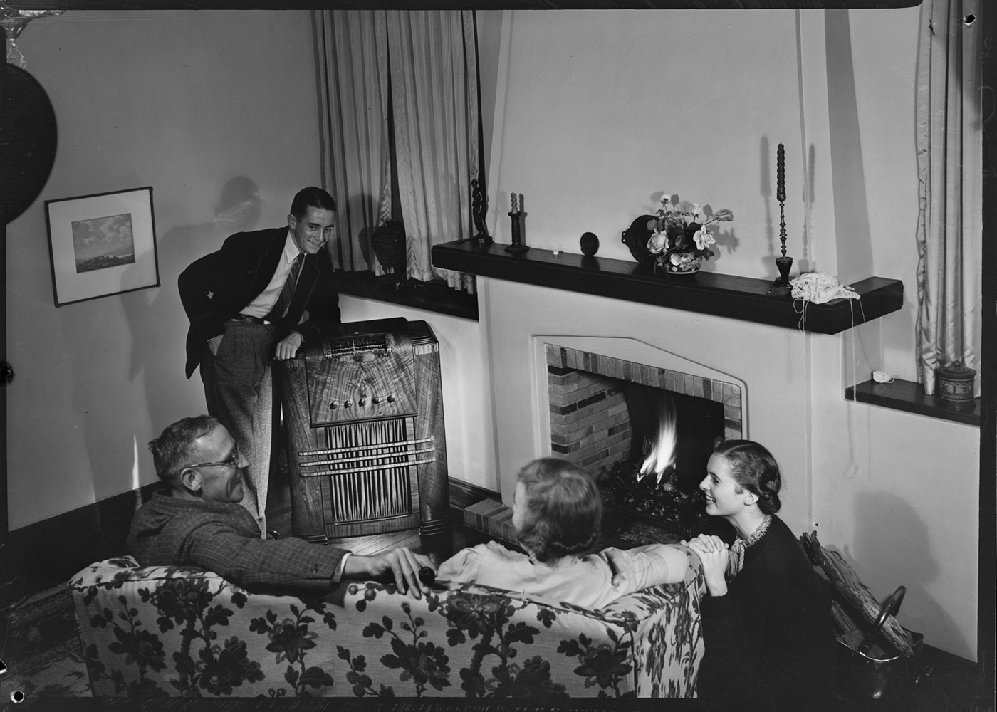 An old black and white photograph three people sitting around a fire place listening to a radio.