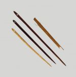 Long wooden needles made of dark brown wood and pointed at the ends with an eye at the wider end.