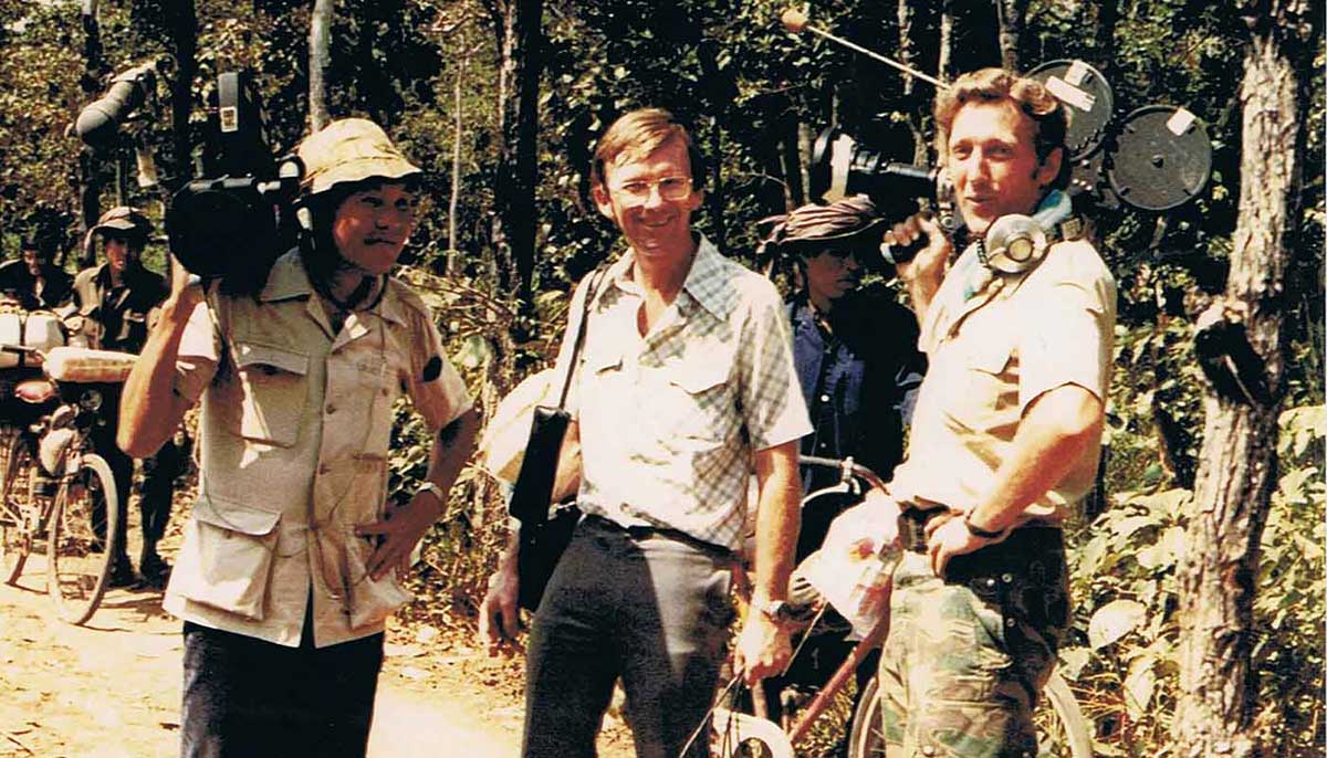 Three men standing on a road. Two rest cameras on their shoulders.