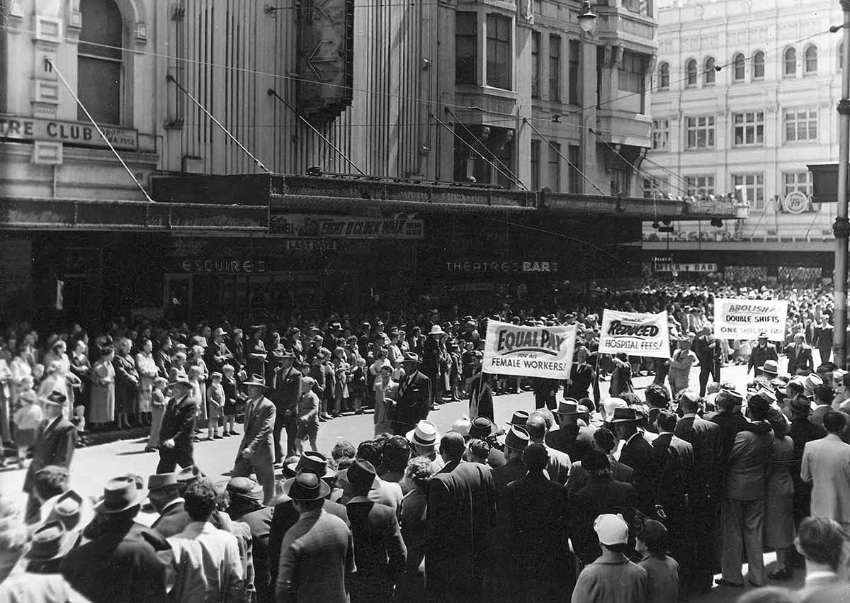 Black and white photo showing men and women marching down street lined with crowds. Workers are holding banners such as Equal pay for female workers.
