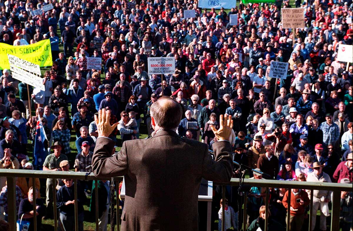 Photo taken from behind Prime Minister Howard who is wearing a bullet-proof vest under his jacket. Facing Howard is a crowd, some of whom are holding placards.