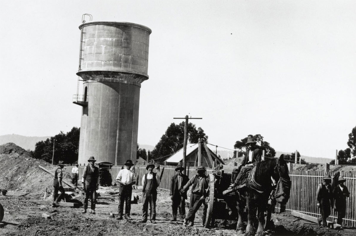 A dozen or so men at work on a suburban construction site, most of them posing for the camera. A draught horse attached to a cart stands among them eating from a nosebag. They appear to be clearing land. A water tower and suburban roofs are visible in the background.