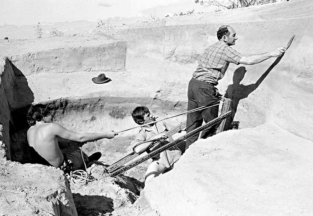 Black and white photograph of three men in a large hole in an arid landscape. One man is scraping at the side of the hole.