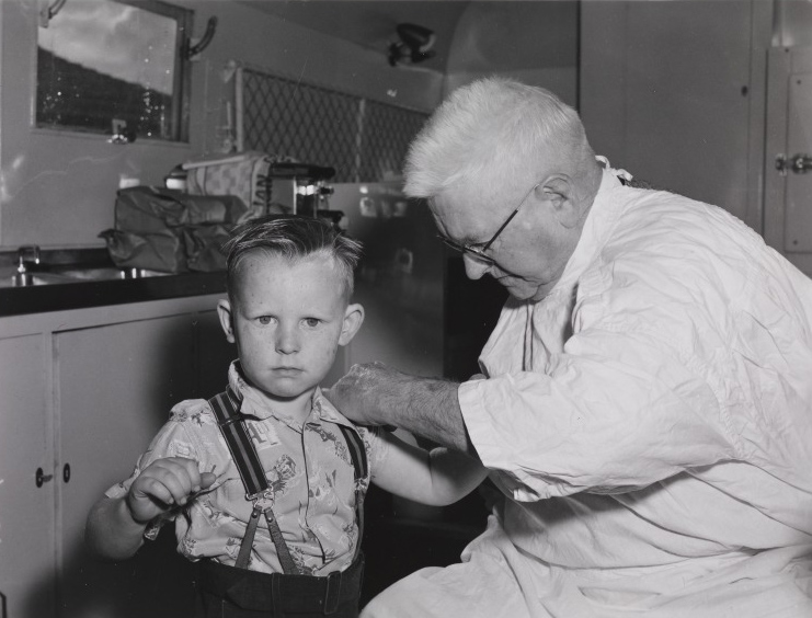 Interior black and white photo of a late middle-aged man in white coat administering injection to the upper arm of a boy, about five