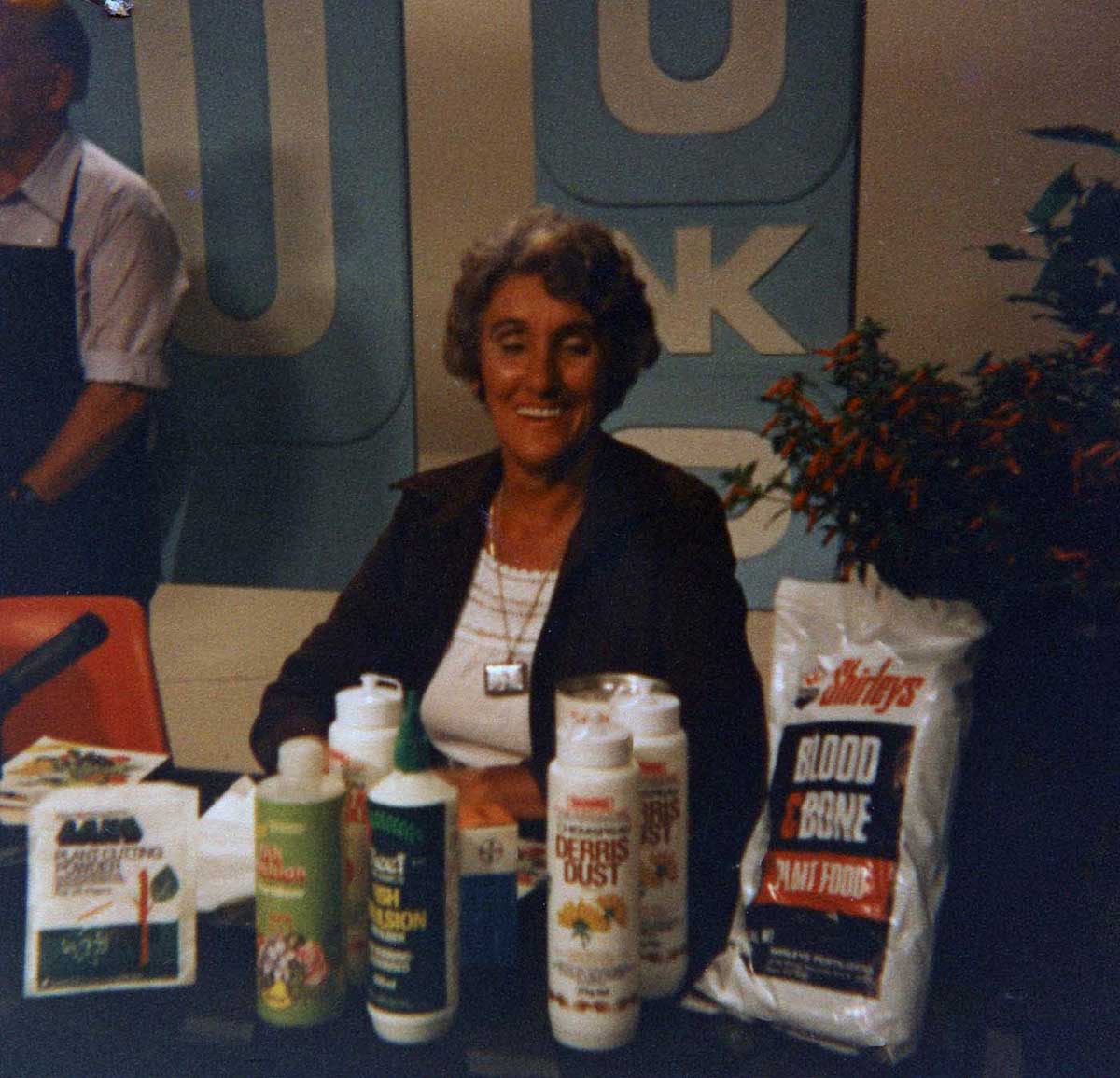 A woman sits at a table laden with gardening items including fertilisers and bottled products. - click to view larger image