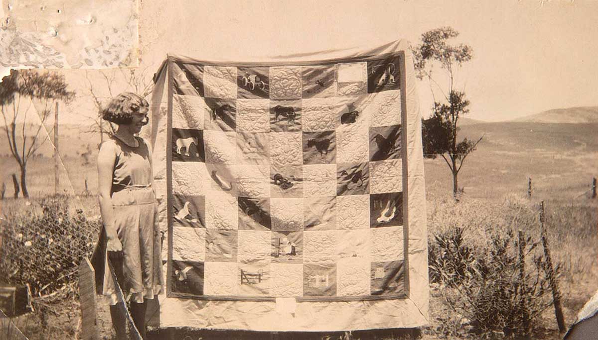 Black and white photo of a young woman with a large quilt. A rural landscape forms the backdrop.