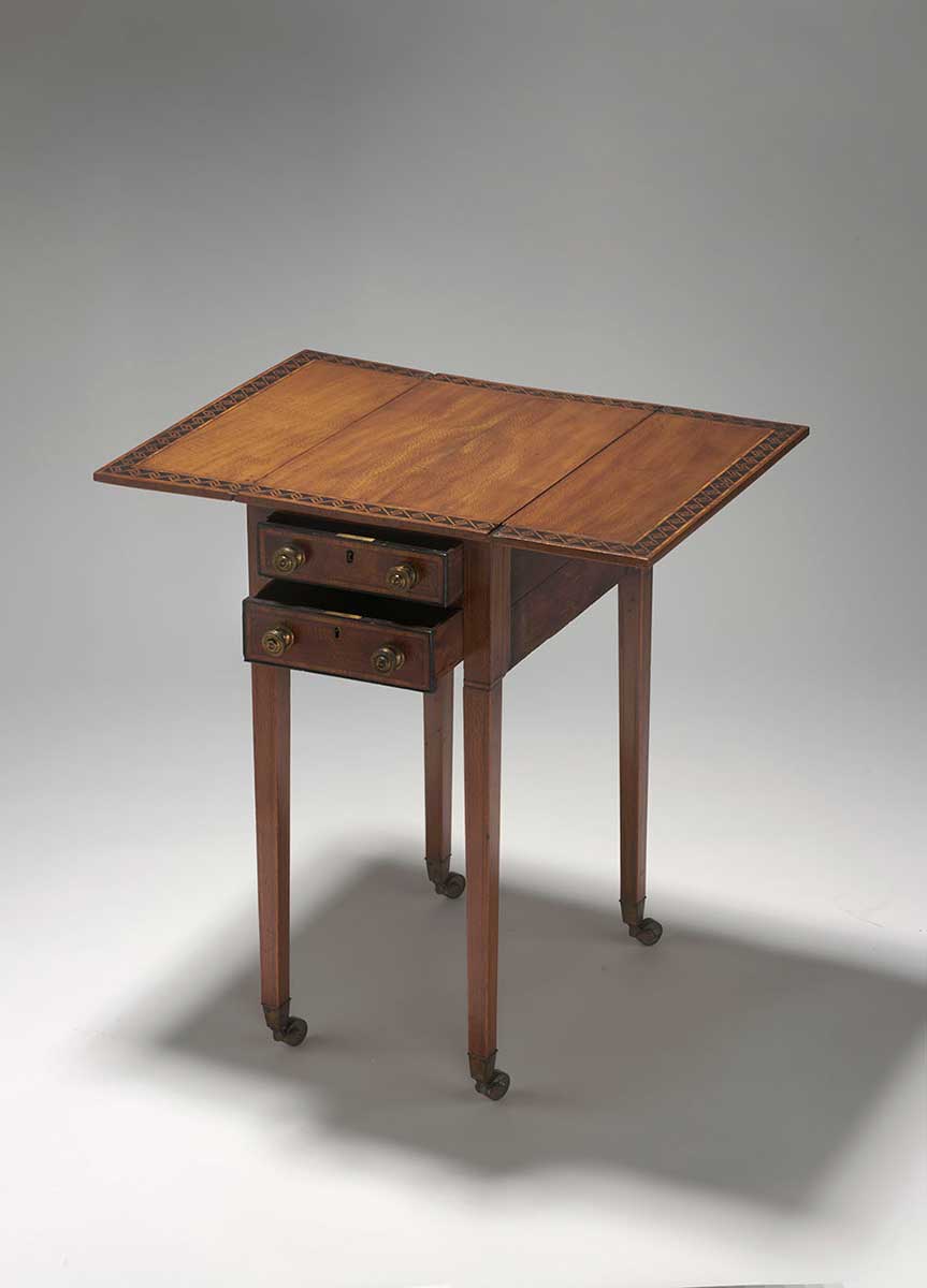 Rectangular wooden table, with fold-down side leaves. The table features narrow inlaid ivory borders, including a wide inlaid border of contrasting timber in a geometric pattern on the table top. The table has four square tapering wooden legs, each terminating in a brass cup and castor. The table has two drawers, each with two round metal knob handles and a brass key-operated lock. - click to view larger image
