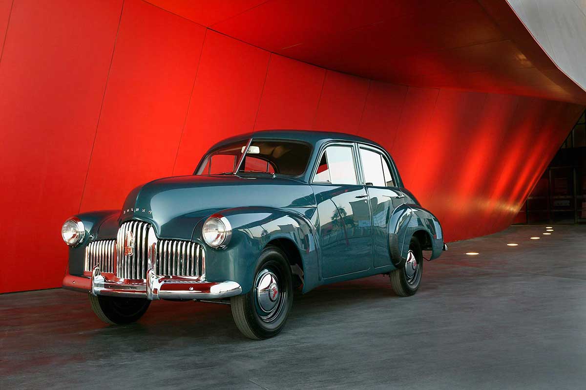 Four door, blue-gunmetal grey sedan with chrome-plated radiator grille, bumper bars, and hub caps. The red entryway to the Museum forms a backdrop.