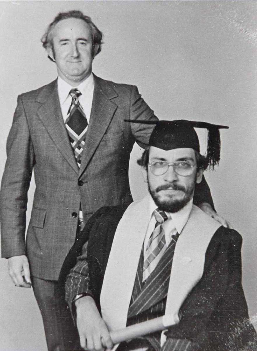 Black and white photo showing a seated man wearing academic gown and hat. A man in s suit stands to his left. - click to view larger image