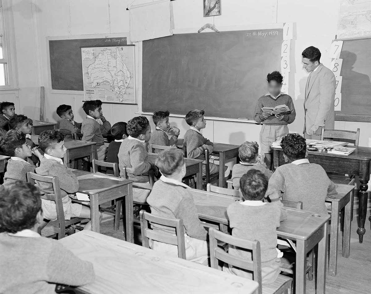 Black and white photo showing young Aboriginal boys in a classroom. One boy stands at the front, reading from a book, with the teacher standing beside him. A map of Australia hangs to the left of a blackboard. A boomerang sits above the blackboard. - click to view larger image