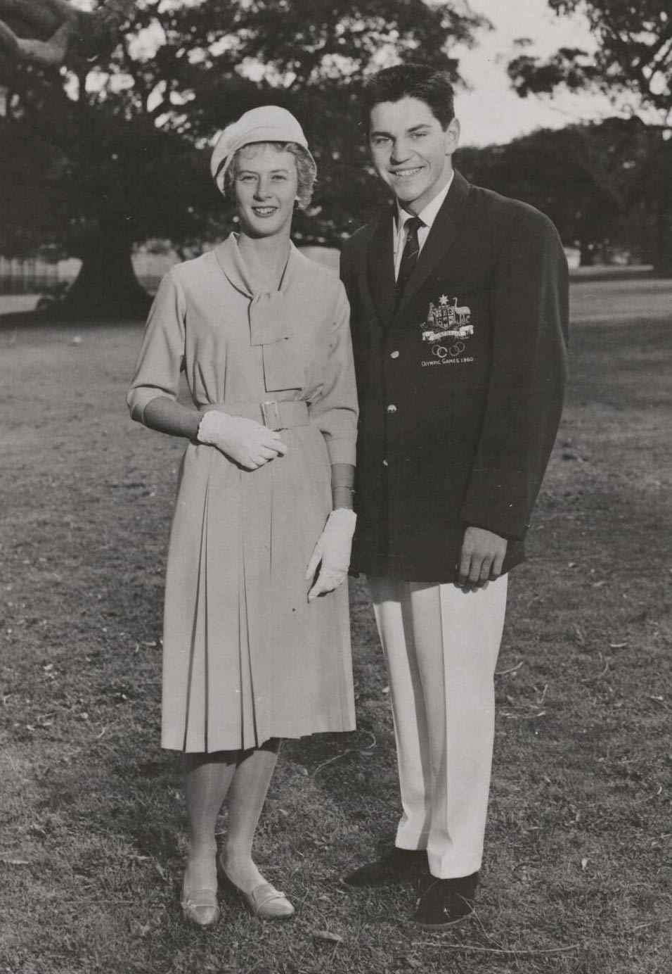 Black and white portrait showing a woman dressed in a knee-length dress and wearing a soft hat, gloves and shoes. A man stands to her right. He wears a dark blazer with an Australian coat of arms on the right breast pocket. He is also wearing a dark necktie, light-coloured trousers and dark shoes. - click to view larger image