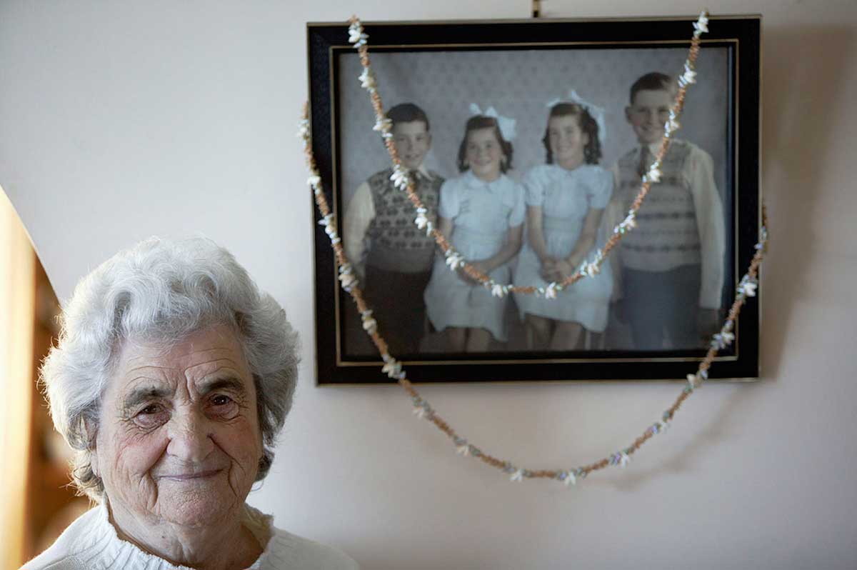 An older woman standing in front of a photograph on a wall. The photograph shows four children. A long necklace made from many small shells is draped over the photograph.