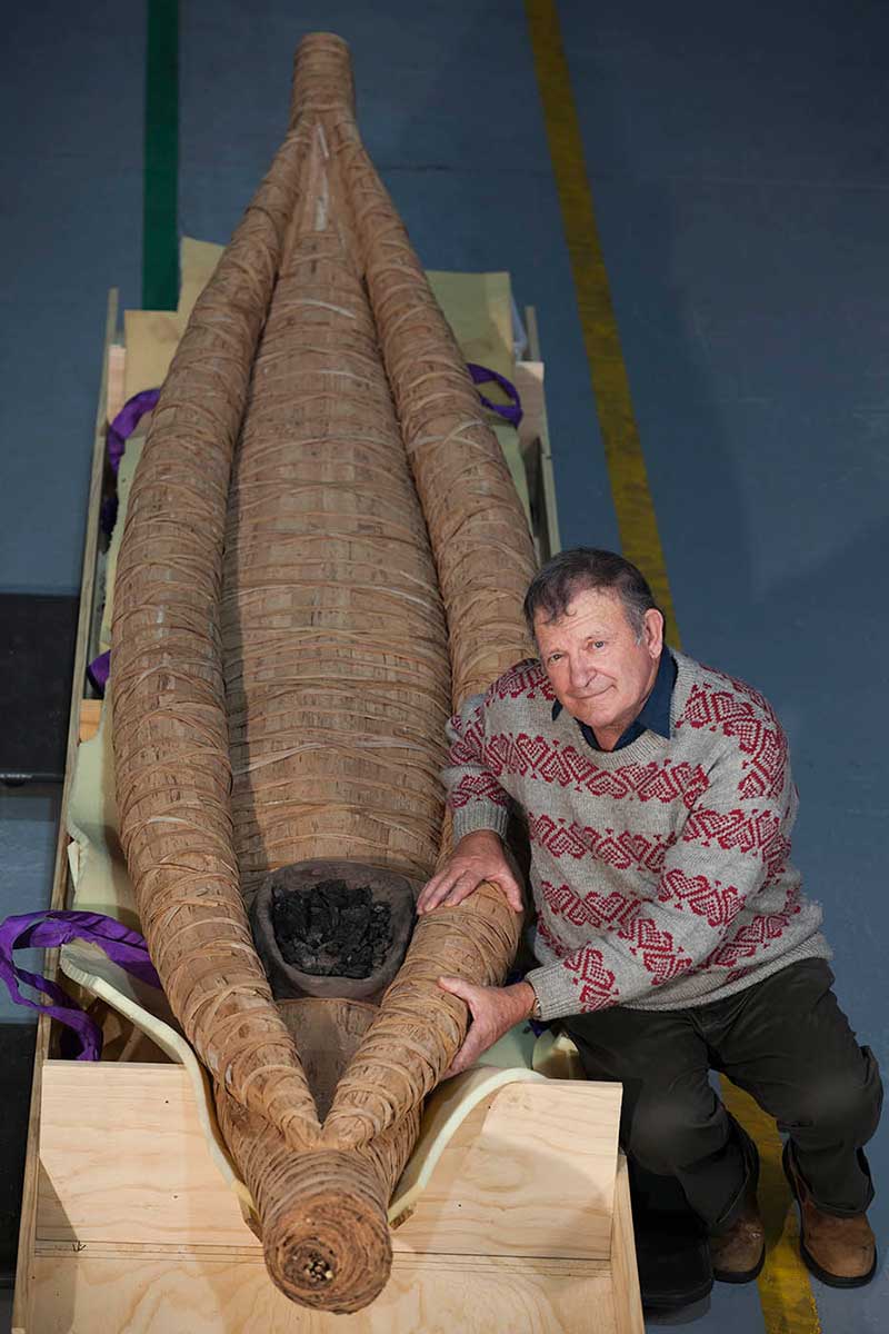 Colour photograph showing a man beside a long bark canoe. A hearth with coals in it sits at the near end of the canoe. - click to view larger image