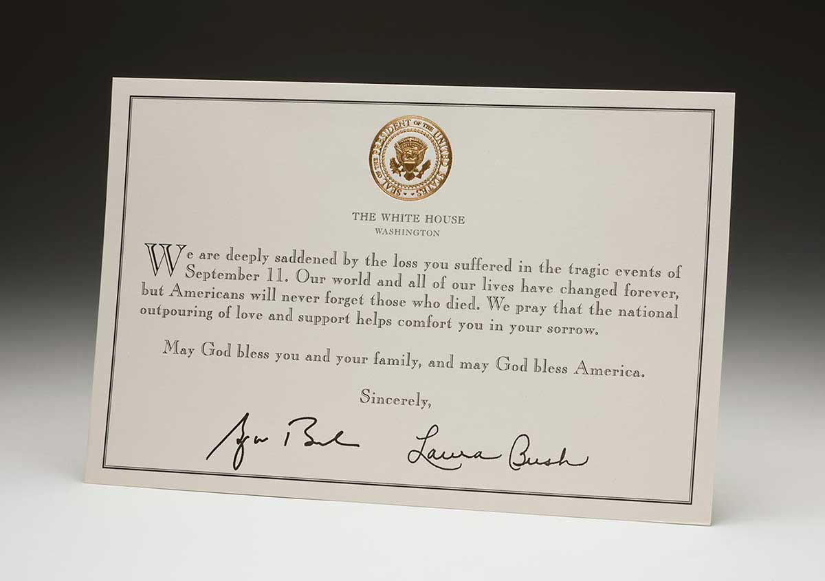 A beige-coloured card with a gold seal at centre top, with printed text 'THE WHITE HOUSE / WASHINGTON'. At the centre is the printed text: 'We are deeply saddened by the loss you suffered in the tragic events of September 11. Our world and all of our lives have changed forever, but Americans will never forget those who died. We pray that the national outpouring of love and support helps comfort you in your sorrow. May God bless you and your family, and may God bless America. Sincerely,'. The card has two handwritten signatures centre bottom, 'George Bush' left and 'Laura Bush' right.