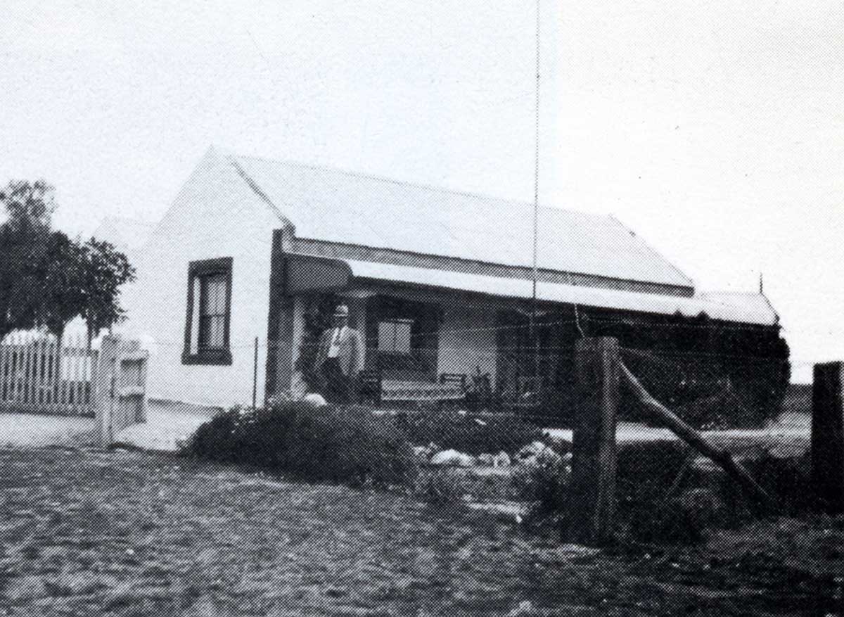 Black and white photo showing a man standing at the front of the small homestead, with a picket fence to the left and a fenced garden at the front. A daybed and wooden chair are on the verandah.