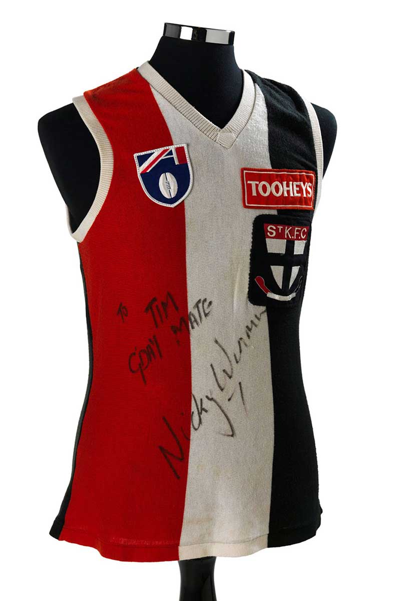 sleeveless top with vertical red, white and black stripes. There is a St Kilda logo on the breast with the word ‘Tooheys’ above that. The words ‘Tim G’day Mate’ is written in texta and signed by Nicky Winmar. - click to view larger image