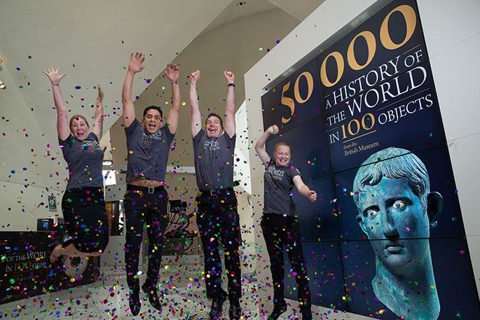 four people jumping for joy alongside a large sign which says '50000 A HISTORY OF THE WORLD IN 100 OBJECTS from the British Museum. A blue-ish grey coloured statue's head is portrayed in the right hand bottom corner. Confetti is being thrown at them.