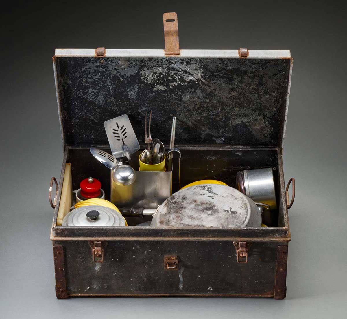 A large metal trunk with a hinged lid. There are two clips at the front and a loop to attach a padlock. The trunk is rusty, has handle on either side and contains cooking utensils. - click to view larger image