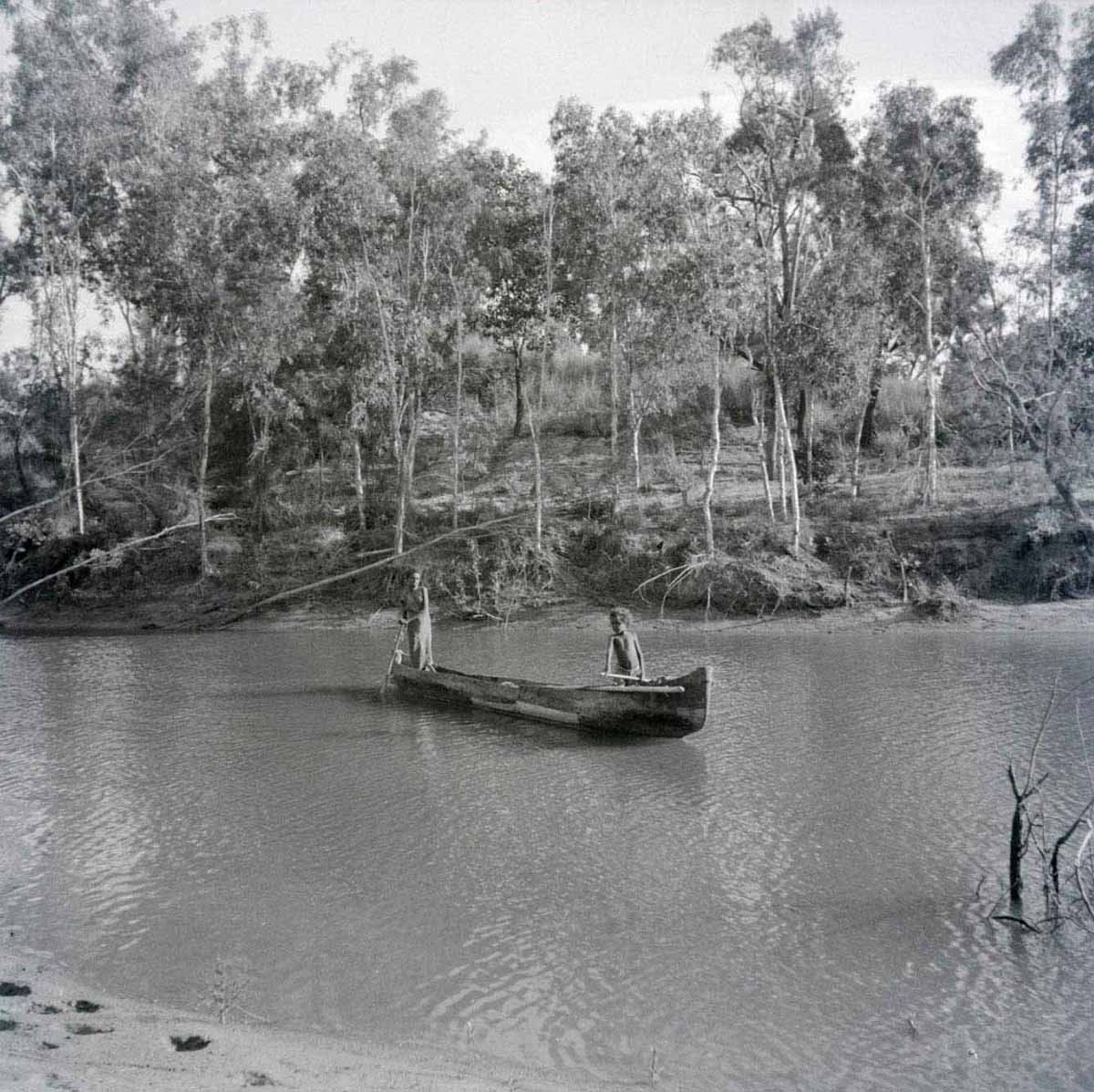 A black and white photographic negative that depicts an Aboriginal child and adult standing in a canoe on a river. - click to view larger image