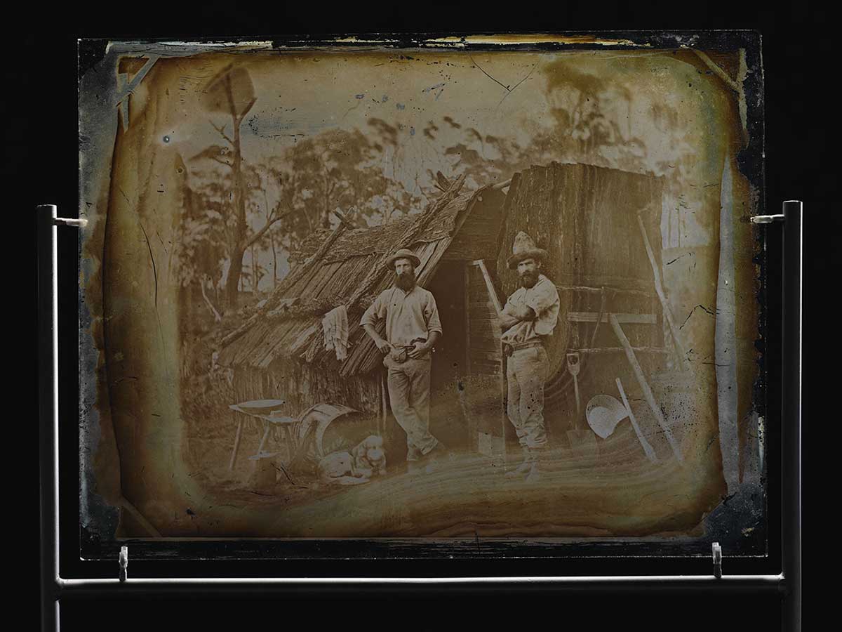 Glass plate showing image of two men standing outside bark slab hut. Both men wear beards and hats. Various tools are propped against the hut and a dog lies to its left.