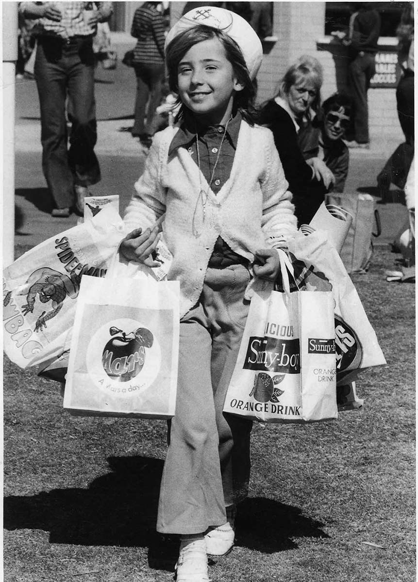 Black and white photo of a child wearing a sailor's hat and pants and carrying multiple show bags on both arms. - click to view larger image