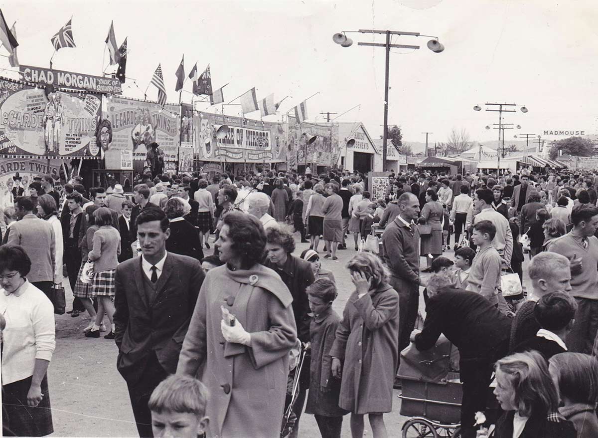 A large crowd of people with many gathering outside tents and buildings with signs advertising 'Chad Morgan', 'Le-Garde Twins' and 'Maze of Mirrors'. Other people are walking down the centre of the alley. - click to view larger image
