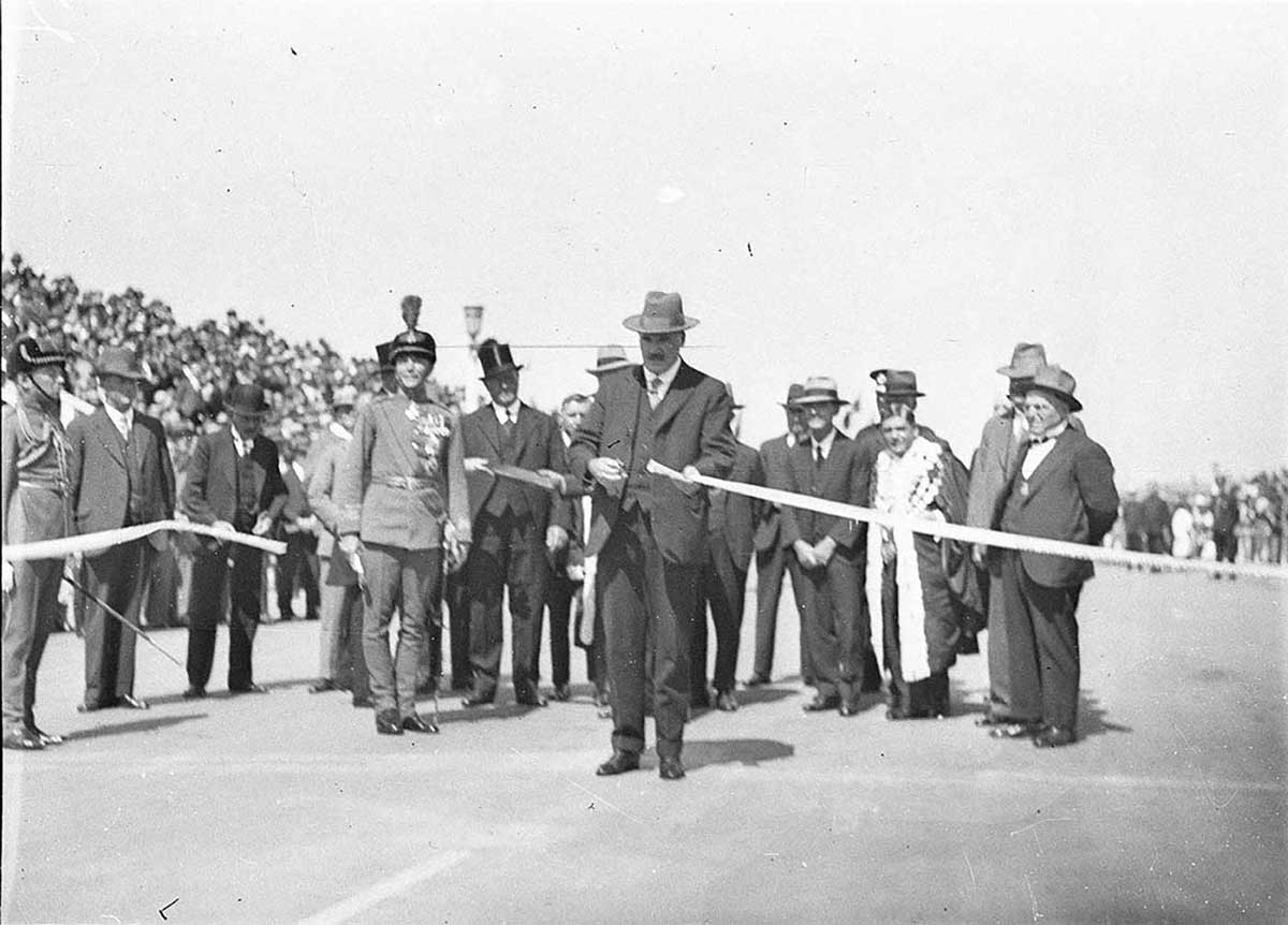 Black and white photo of dignitaries, with one man standing forward to cut a ribbon. - click to view larger image