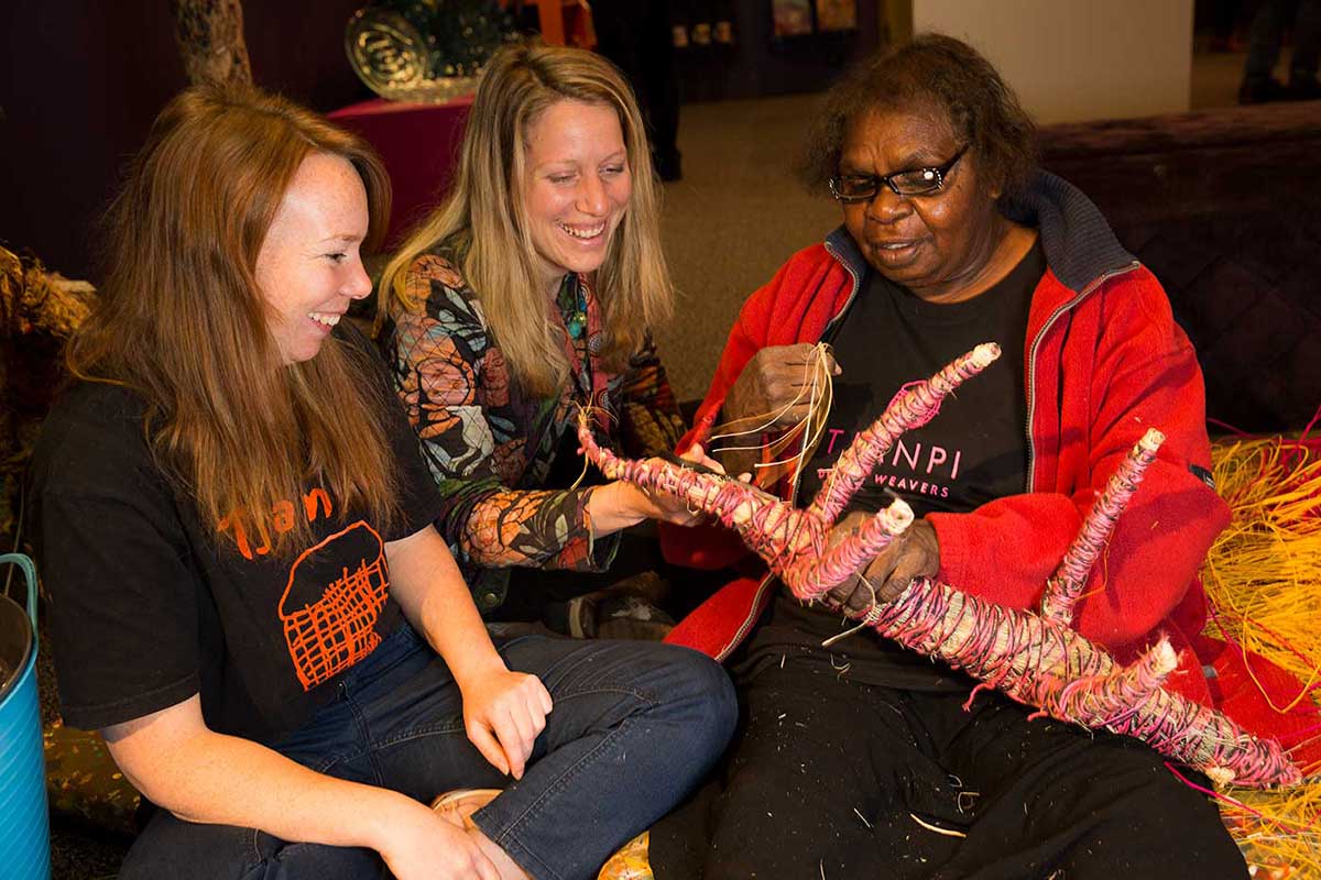 Three women sitting and discussing woven object. - click to view larger image