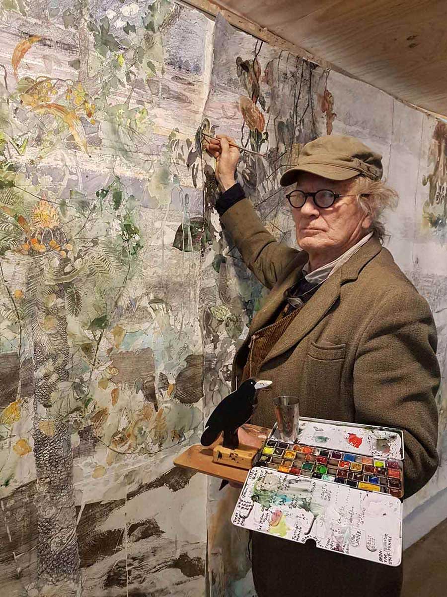 John Wolseley standing in front of a large artwork with his brush raised to the paper and a palette in the other hand. - click to view larger image