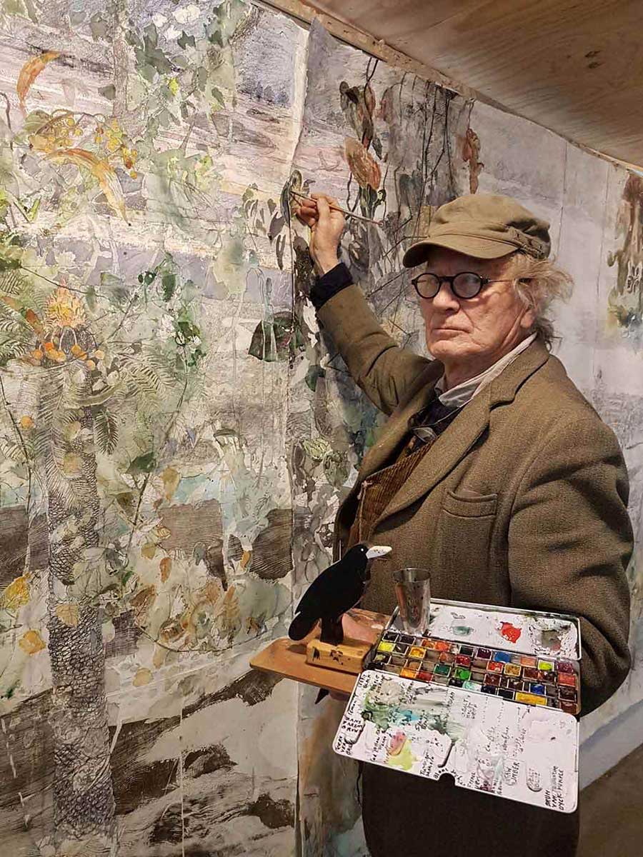 The artist stands in front of a large artwork with his brush raised to the paper and a palette in the other hand. - click to view larger image