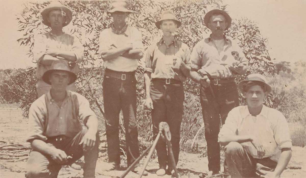 Six men – four standing, two crouching – wearing similar clothing, namely black pants, light shirts and broad-brimmed hats.