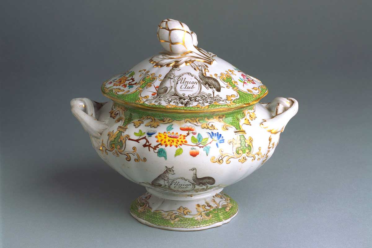 Image showing a ceramic soup tureen has a white base and gold-rimmed lid and handles. It is decorated with elaborate imagery including flora and curlicules. A black and white kangaroo and emu appear either side of a stylised 'Union Club' coat of arms.