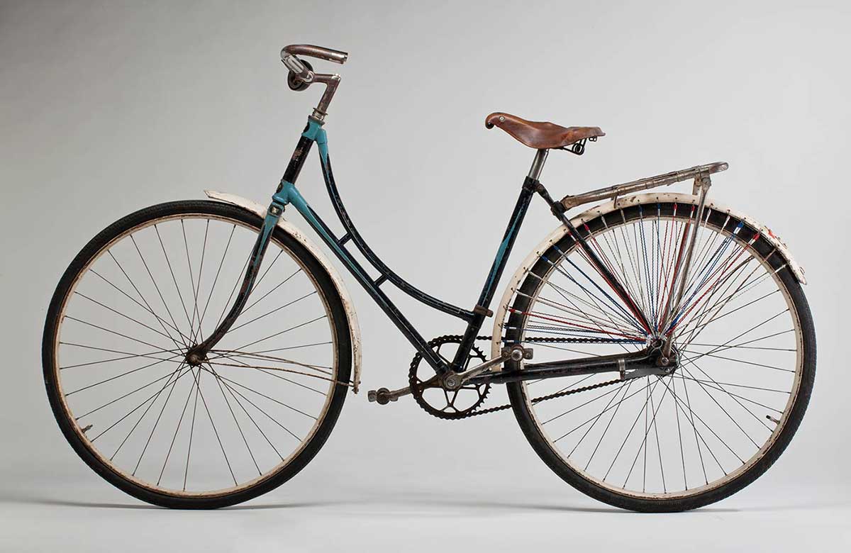 Ladies refurbished 'Roadmaster' bicycle. The frame is handpainted in turquoise and navy blue enamel paint, and the mudguards are white. It has a brown leather seat and a silver rack attached to the rear end. Both of the wheels are from a Malvern Star. The rear wheel has lengths of red, white and blue fine cord attached through the mudguard and gathering just above the centre of the wheel; on both sides of the rear wheel. Small reflector on rear wheel guard. Large black bell on right side of the handlebar.