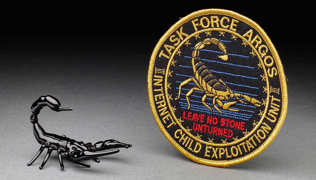 A small black glass scorpion with reared tail beside  circular path showing a scorpion and the words 'Task Force Argos Internet Child Exploitation/Leave no stone unturned'. - click to view larger image