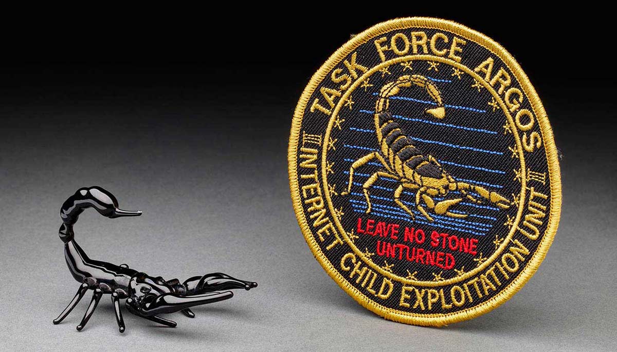 A small black glass scorpion with reared tail beside circular path showing a scorpion and the words 'Task Force Argos Internet Child Exploitation/Leave no stone unturned'. - click to view larger image