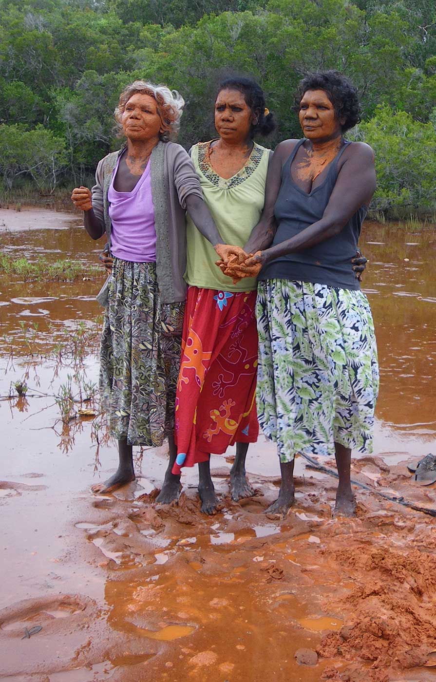 Three ladies in stand bare-footed in red clay. Their hands and faces are painted with clay. - click to view larger image