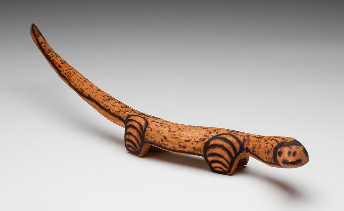 Thin black and brown wooden sculpture of a goanna, standing upright on three of four small short legs. The head and neck are upright and pointing slightly to the right. The thin tail points straight out and curves to the left. The top exterior is covered with burnt engraved designs, and the small pointed head has burnt engraved eyes, nostrils and mouth. Two thick black band arcs meet in the centre above the eyes. The neck, body and long tail are covered with a lightly shaded speckled pattern. The four short legs are covered with parallel horizontal arcs. - click to view larger image