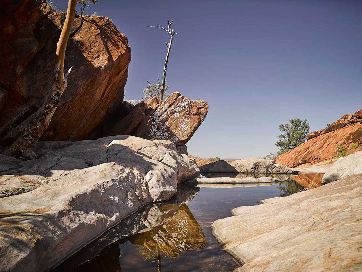 Colour photograph showing a rocky landscape with a pool of water at the centre. A large reddish rock at the left has designs painted in white. - click to view larger image