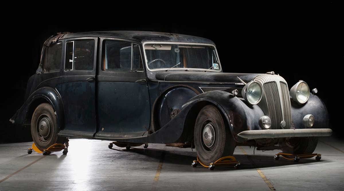 A long, sedan-style car with faded black paint, on jacks in an exhibition space. - click to view larger image